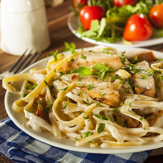 Close up of a white plate full of Fettuccine Alfredo with slices of grilled chicken, garnished with fresh green herbs.
