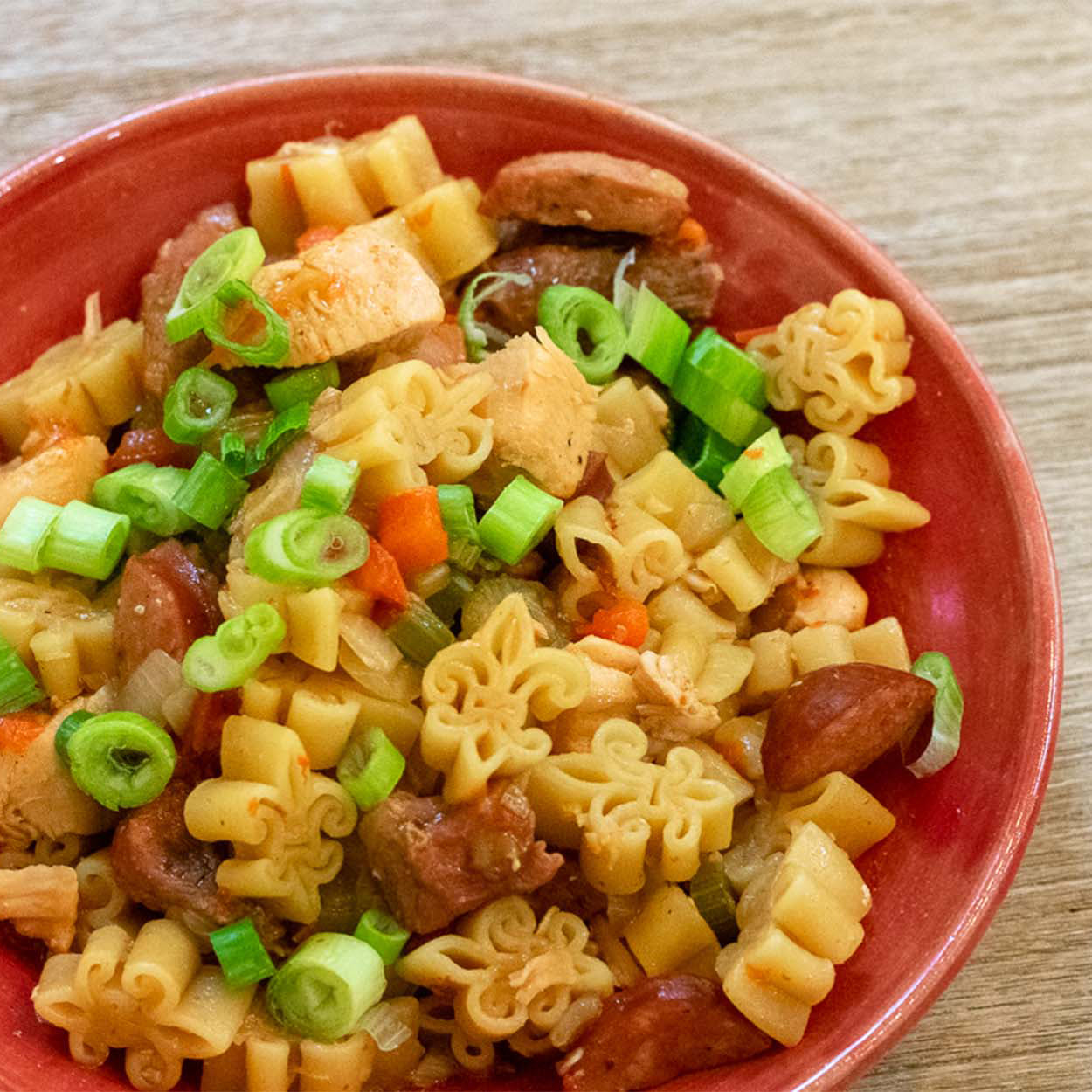 Close up of a red bowl filled with pastalaya, consisting of fleur de lis shaped pasta with andouille sausage, chicken, and vegetables.