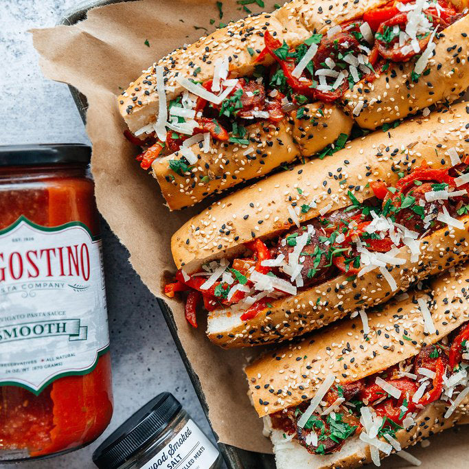 Close up of a partially obscured jar of Dagostino Smooth Tomato Sauce next to a platter of Italian sandwiches on seeded bread.