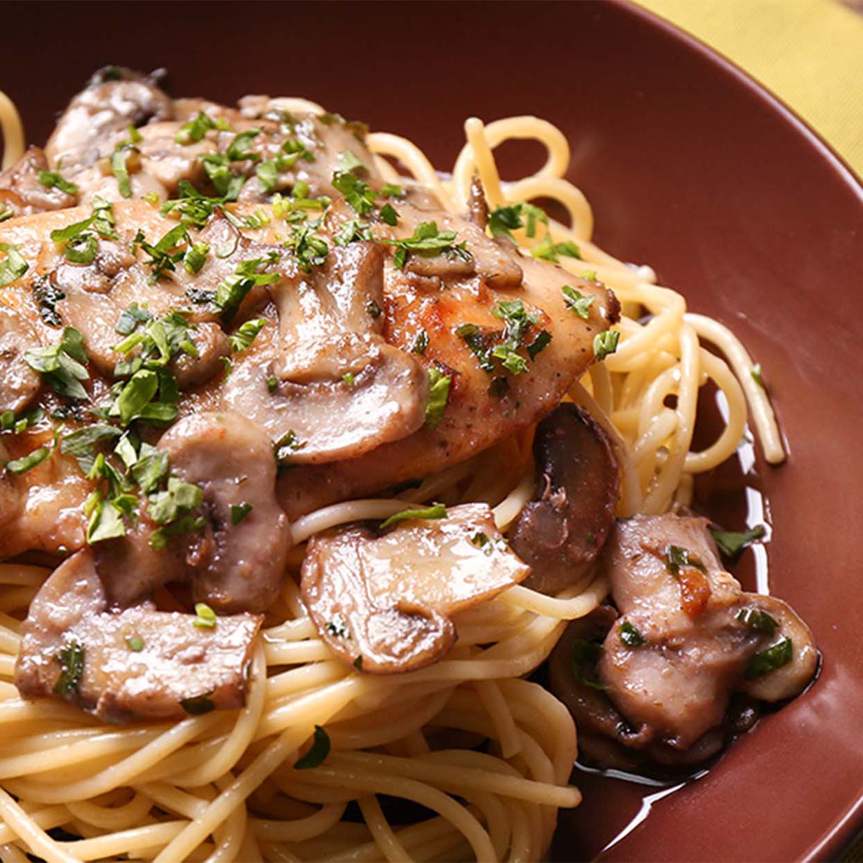 Close up of a dark brown plate filled with chicken marsala on cooked spaghetti